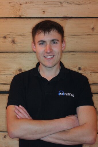 Nico Japes, professioneller Hochzeits-DJ und Inhaber von NJ-Eventing aus Kirchhundem. Er lächelt im Marken-Shirt vor einer Holzwand. Ihr Experte für Eventtechnik im Kreis Olpe und Sauerland.